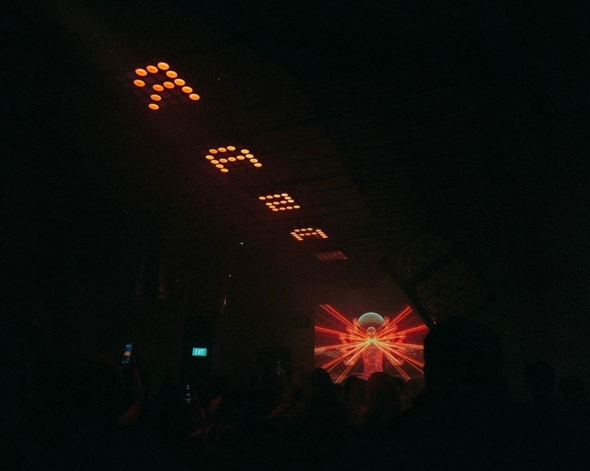 Square LED panels glowing orange along a dark ceiling above a crowd, with a red visual on a screen.