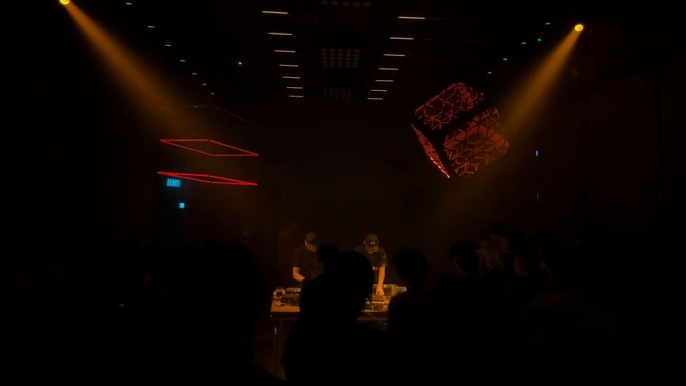 Two amber beams crossing above the DJs, with red neon frames and ceiling light bars.